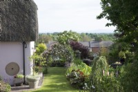 Vue sur jardin avec arche de clématites et chaumière surplombant les maisons environnantes et la campagne début juin.