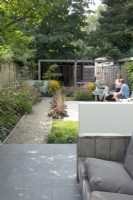 Jardin mitoyen aux murs bas blancs. Vue d'ensemble jardin avec pergola, terrasse, allée, bancs en bois.