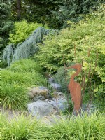 Sculpture en métal de héron et de joncs dans un parterre de fleurs à sec avec feuillage d'hémérocalles, noisetier d'hiver et cèdre de l'Atlas bleu pleureur
