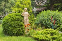 Sculpture de femme près de conifères : Juniperus chinensis 'Old Gold' - Genévrier de Chine, Thuja occidentalis 'Little Giant' - Cèdre en parterre de fleurs en été, Québec, Canada - Juillet