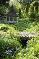 Simple pont en bois sur un ruisseau à l'ancien presbytère, Netherbury, Dorset en mai bordé de primevères, iris et fougères et un pavillon en bois derrière