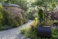 Un chemin de gravier mène à travers un jardin d'été avec un mur en torchis soutenant un parterre de fleurs. Rouleau à gazon en premier plan.