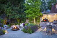 Jardin en contrebas illuminé et coin repas extérieur avec table en bois, chaises et unité de cuisine sur mesure avec four à pizza intégré et barbecue la nuit.