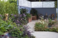 Un sanctuaire serein avec un coin salon couvert à côté d'un étang planté d'Iris 'White Swirl', et entouré de plantations herbacées dans les tons de vert, violet, bronze et bleu.