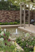 Un bassin en contrebas avec jets d'eau est bordé de parterres herbacés plantés d'une tapisserie de lupins, alliums, geums, pimpinella, verbascums et graminées ornementales.