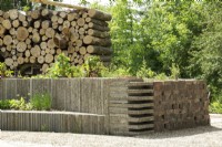 Banc et pots de pavés recyclés et drainage fait de vieilles cuillères en fer. Mur de troncs d'arbres empilés et de tuyaux de drainage en béton.