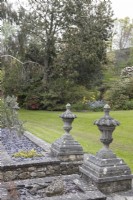 Un large mur de soutènement avec des embouts en pierre formels mène à une pelouse avec divers arbres et arbustes en arrière-plan. Ferme de Whitstone, jardin NGS Devon. Le printemps.