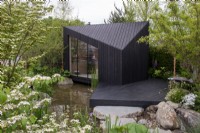 Une cabane en mélèze carbonisé avec terrasse en bois surplombe un étang entouré de Betula nigra - A Garden Sanctuary by Hamptons, RHS Chelsea Flower Show 2022