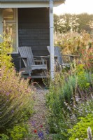Chemin de coquillages entre parterres de fleurs avec vivaces et graminées jusqu'au porche ouvert avec sièges en bois.