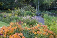 Chemin de terrasse en bois menant à des sièges à côté d'un parterre de fleurs avec des Alstromerias orange, des Cordylines et des Astilbes