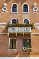 Balcon en pierre ornementale altérée avec des jardinières de pélargoniums rouges devant des fenêtres à volets verts et un mur en stuc marron.
