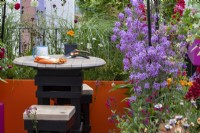 Une table de jardin en bois et deux tabourets entourés d'Hesperis matronalis, de Salvia 'Bordeaux', d'Eschscholzia californica et d'ornements gazonnés sur .Le Cirrus Garden - designer : Jason Williams