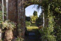 Une vue à travers une arche dans une brique rouge, un vieux mur d'un chemin couvert de mousse au-delà et diverses plantes qui poussent autour. Regency House, jardin Devon NGS. L'automne