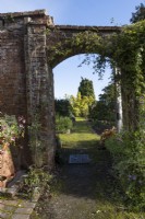 Une vue à travers une arche dans une brique rouge, un vieux mur d'un chemin couvert de mousse au-delà et diverses plantes qui poussent autour. Maison Regnecy, jardin Devon NGS. L'automne