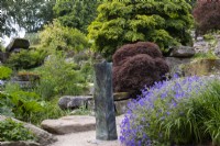 Une sculpture en métal entourée de géranium, d'arbres et de formations rocheuses à Paxton's Rock Garden, Chatsworth House and Garden.