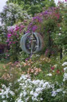 Une sculpture de girouette s'élève au-dessus des roses 'Flower Carpet White' et 'Flower Carpet Amber'. Derrière, sur la pergola, la rose 'Veilchenblau', 'Sir Paul Smith' ('Beapaul') et 'Paul's Himalayan Musk'.