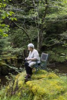 Visiteur assis sur une chaise sur un monticule couvert de mousse par stream holding camera. Peut.