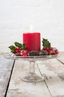 Décoration de table de Noël avec bougie rouge, cônes et feuillage Ilex et baies sur présentoir à gâteaux en verre vintage