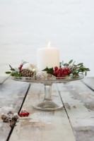 Décoration de table de Noël avec bougie blanche, cônes, boules et baies de feuillage Ilex sur présentoir à gâteaux en verre vintage