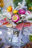 Bouquet de fleurs contenant de la paille, du statice et des graminées dans un vase en verre décoré de feuilles automnales.