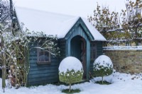 Bâtiment de jardin en bois peint vert foncé en hiver avec paire d'arbres topiaires en face de l'entrée. Décembre.