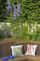 Coin salon en contrebas bordé d'un parterre de fleurs ensoleillé planté de delphiniums, de lupins et d'iris dans le New Blue Peter Garden - Discover Soil, RHS Chelsea Flower Show 2022