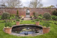 Vue sur jardin clos formel avec porte gothique avec petite piscine circulaire bordée de pots en terre cuite de tulipes bordeaux sous-plantées d'altos jaunes