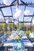 Table dressée dans une serre avec des assiettes, des couverts, des verres, des bougies et un présentoir de fleurs au milieu et des pompons en papier suspendus