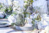 Table dressée dans une serre avec assiettes, couverts, verres, bougies et un présentoir de fleurs au milieu avec des décorations de Pâques