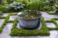 Une piscine d'eau galvanisée placée sur des briques étroites avec Hydrocotyle tripartita sp. 'Japon' poussant entre les pavés. Myeloma UK - A Life Worth Living Garden, gagnant de l'or. Créateur : Chris Beardshaw