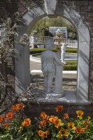 Une statue en pierre, de dos, d'une personne nue appuyée sur un piédestal. Tulipes en fleur devant et la statue est placée dans une arche dans un mur de briques avec des boutons de fleurs de glycine autour de l'arche. Trago Mills montrent les jardins, Devon, Royaume-Uni. Peut. Printemps