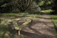Un banc en bois triangulaire au bord d'un chemin dans un jardin boisé. Jardins de Marwood Hill, Devon. Printemps. Peut.
