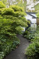 Une allée en blocs de chêne bordée d'érables et de vivaces feuillues mène à un bâtiment de jardin.