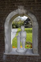 Statue en pierre d'un personnage debout avec un bras autour d'un buste sur un piédestal, installée dans une arche dans un mur de briques avec vue sur des pelouses et un bâtiment à pans de bois. Trago Mills montrent les jardins, Devon, Royaume-Uni. Peut. Printemps