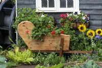À côté d'une ancienne roue à aubes, une jardinière de brouette est fabriquée à partir d'une ancienne table à manger et remplie de pélargonium traînant.