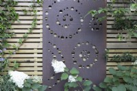 Écran métallique avec un design circulaire et en spirale attaché à une clôture en bois contemporaine, hortensia à fleurs blanches au premier plan