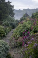 Chemin pavé menant à travers des parterres de fleurs d'automne brumeux dans un jardin de style cottage, maison d'été carrelée derrière