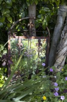 Un miroir aux bords en métal rouillé niché à côté d’un petit tronc d’arbre parmi une variété de feuillage. Juin.