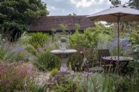 Petit jardin de cour rempli de plantations de fin d'été, avec bâtiment en chêne à l'arrière et pièce d'eau, bain d'oiseaux et table avec parasol au premier plan
