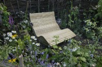 Une chaise en bois incurvée à côté de plantes vivaces d'été enfermées dans une spirale de haie morte en brosse de bouleau tissée conçue par RHS Garden Wisley exposée au Hampton court Palace Garden Festival 2023.