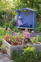 Potager avec semis et outils exposés sur parterre de fleurs surélevé. En arrière-plan, une femme dans un petit belvédère se repose du travail.