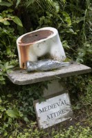 Un vieux torse de mannequin repose sur un socle avec une bouteille en verre en forme de poisson. Un panneau indiquant « Tenue médiévale » se trouve en dessous. Le puzzle sculpture représente un morceau de morue. Lumières du port, jardin Devon NGS. Juillet.