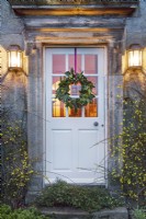 Couronne de Noël de laurier portugais et de cornouiller accrochée à la porte de la maison. Lampes d'entraîneur allumées et jasmin à fleurs d'hiver de chaque côté de la porte.