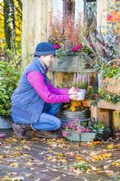 Femme plaçant un petit pot de Callunas sur un banc avec un panier en osier. Feuilles éparpillées sur une terrasse en bois