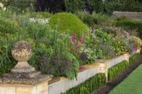 Un muret avec un fleuron rond en pierre conserve le parterre de fleurs surélevé du Bourton House Garden, Gloucestershire. Contre le mur se trouve une haie basse d'Euonymus japonicus 'Green Spire''.