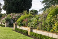 Un muret avec un fleuron rond en pierre conserve le parterre de fleurs surélevé du Bourton House Garden, Gloucestershire. Contre le mur se trouve une haie basse d'Euonymus japonicus 'Green Spire''.