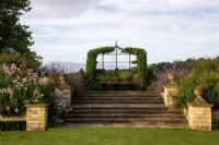 De larges marches et des piliers en pierre surmontés de fleurons ronds mènent à un siège sous une tonnelle recouverte de lierre sur la promenade surélevée du Bourton House Garden, Gloucestershire.