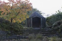 Des marches mènent à une maison d'été rustique en bois avec un Prunus Taihaku à gauche avec un feuillage d'automne. La maison du jardin, Yelverton. Automne, novembre