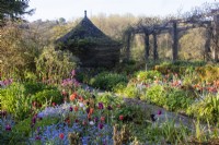 Soleil matinal sur des parterres de fleurs printaniers remplis de tulipes et de myosotis en regardant vers une maison d'été carrelée et une pergola au Gravetye Manor.