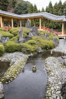 Vue sur zone pavée avec petits bassins et rochers placés avec parterre de rhododendrons (pas en fleur). Marches et pavillon du bâtiment du temple.